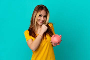 Teenager blonde girl holding a piggybank over isolated blue background celebrating a victory
