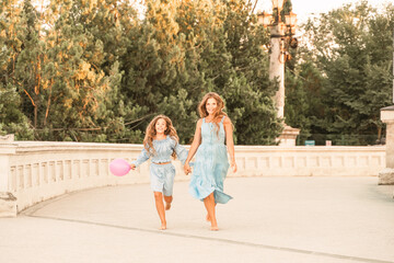 Daughter mother run holding hands. In blue dresses with flowing long hair against the backdrop of a sunset and a white building.