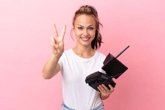 Teenager Russian girl holding a drone remote control isolated on pink background smiling and showing victory sign
