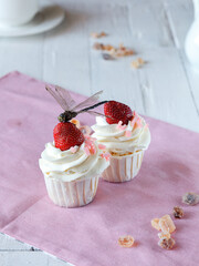 two cupcakes with white cream and strawberries and dragonfly on a light wooden table