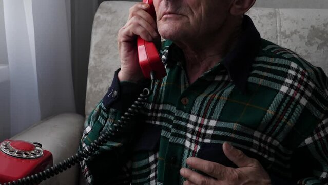 a pensioner holding his heart calls calling for medical help while at home. an elderly man holds his hand to his chest and calls on a wired vintage phone