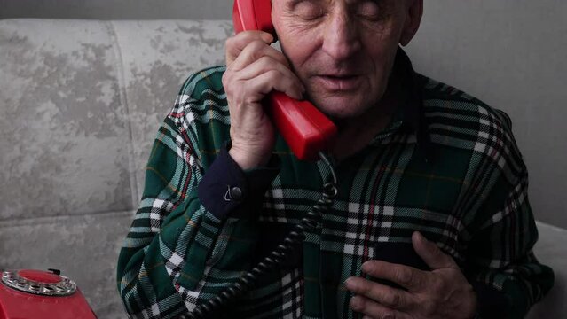 a pensioner holding his heart is calling for medical assistance while sitting in a chair. an elderly man holds his hand to his chest and calls on a wired vintage phone