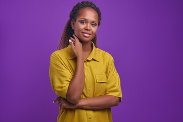 Young beautiful optimistic African American woman millennial in yellow casual shirt touches neck and slightly smiling looks at camera stands on isolated purple background. Ethnic girl