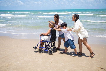 Happy holidays with family. Travel and vacations concept. Happy Asian family and woman elderly handicap on wheelchair walking together on the beach.