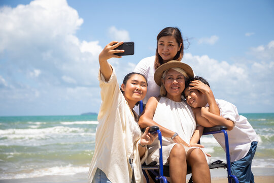 Happy holidays with family. Travel and vacations concept. Happy Asian family and woman elderly handicap on wheelchair making salfie together on the beach.