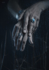 Women's hands with jewelry, photographed through fogged glass with water droplets. Mystical forms. Dark fantasy. Fashion. Body parts. Exquisite fingers.