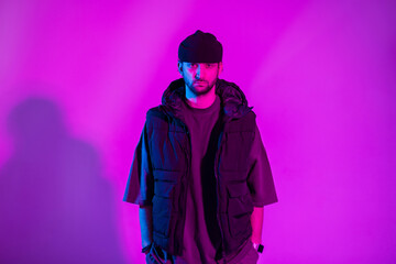 Fashion trendy professional dancer man with hat and stylish vest and t-shirt stands in colorful studio with pink and neon light