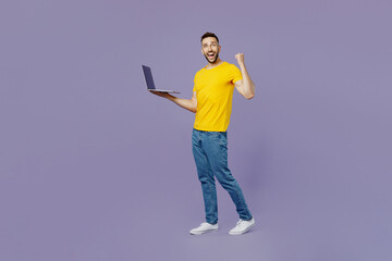 Full body side view fun young man wear yellow t-shirt hold use work on laptop pc computer do winner gesture walk go isolated on plain pastel light purple background studio portrait. Lifestyle concept.