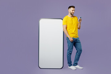 Full body side view young caucasian man wears yellow t-shirt near big huge blank screen mobile cell phone with area use smartphone browsing internet isolated on plain pastel light purple background.