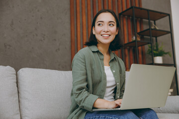 Young IT woman of Asian ethnicity wear casual clothes work hold use laptop pc computer look aside sit on grey sofa couch stay at home hotel flat rest relax spend free spare time in living room indoor