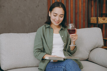 Obraz premium Young calm smiling smart woman of Asian ethnicity wears casual clothes read book drink red wine sits on grey sofa couch stay at home hotel flat rest relax spend free spare time in living room indoor.