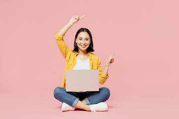 Full body fun young IT woman of Asian ethnicity wear yellow shirt white t-shirt sitting hold use work on laptop pc computer point finger aside on area isolated on plain pastel light pink background.