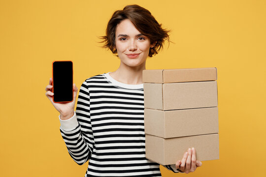 Young woman wearing casual striped shirt holding in hand stack cardboard blank boxes use mobile cell phone with blank screen workspace area isolated on plain yellow color background studio portrait.