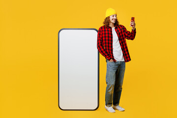 Full body smiling young man wearing red checkered shirt white t-shirt hat big huge blank screen mobile cell phone with area using hold smartphone isolated on plain yellow background studio portrait.