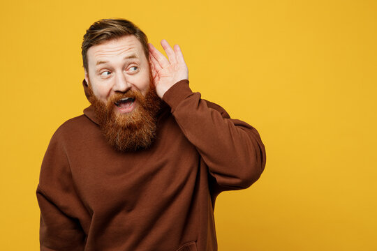 Young curious nosy smiling redhead caucasian man wearing brown hoody casual clothes try to hear you overhear listening intently isolated on plain yellow background studio portrait. Lifestyle concept.