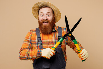 Happy fun young bearded man wears straw hat overalls work in garden hold in hand hedge shears scissorries isolated on plain pastel light beige color background studio portrait. Plant caring concept.