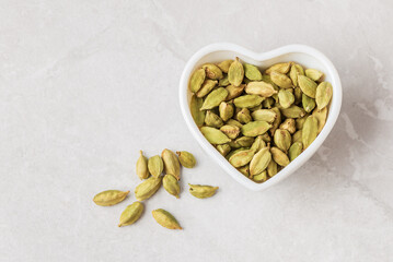Aromatic condiment cardamom pods with seeds in white heart shape bowl  and scattered on light backdrop top view with copy space