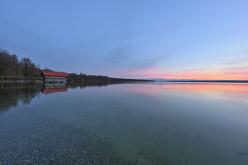 Ein Bootshaus steht im See und im Hintergrund sieht man die Berge im Abendrot.