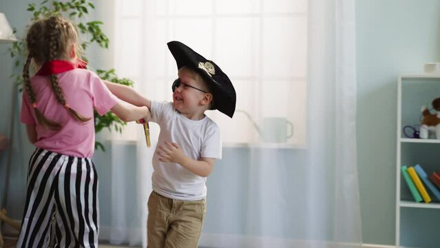 Toddler boy in pirate hat and eye patch attacks elder sister with toy sword playfully. Happy children siblings play duel running around room and making noise