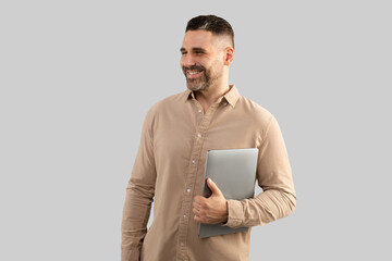 Happy middle aged caucasian male entrepreneur holding laptop, looking aside at free space and smiling, grey background