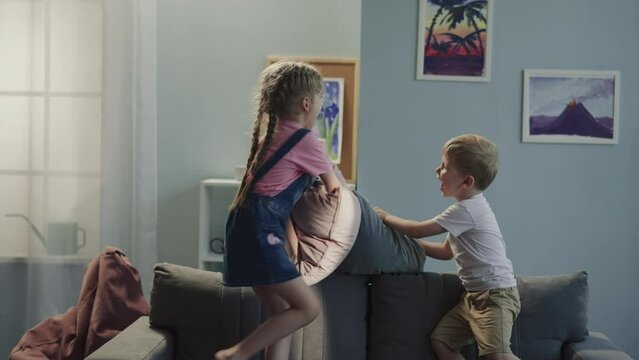 Laughing sibling children have pillow fight jumping on soft sofa at home. Active little boy and preschooler girl play together in light living room