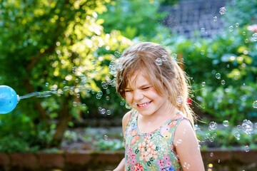 Happy little blonde preschool girl having fun with blowing soap bubble blower. Cute child playing...