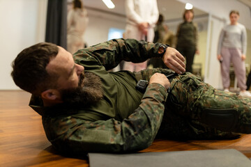 Fototapeta premium A bearded man in his 40s, a military medic lies on the floor and demonstrates to civilians in the room how to apply a combat medical tourniquet to a leg