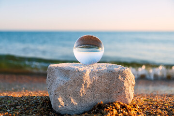 Glass lens globe on sandy beach on sea surf and blue sky background at sunset.