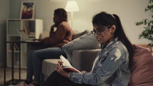 Indian woman reads book sitting in bean bag chair while African American girl friend studying on laptop. Students prepare for classes in college at home