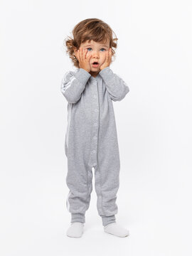 A toddler 1-2 years old expresses horror, disappointment, sadness. He clasped his hands around his face in a gray jumpsuit on a white background.