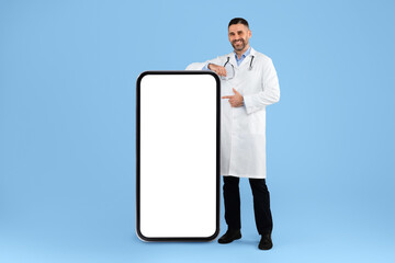 Male physician in white uniform pointing finger at huge cellphone with blank screen on blue studio...