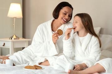 Cheerful european millennial woman and little girl in bathrobes sit on bed, enjoy weekend together, drink coffee