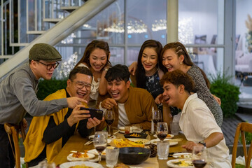 Group of Asian people using mobile phone taking selfie together during outdoor celebration dinner party in the garden on summer holiday vacation. Man and woman friend reunion meeting at restaurant.
