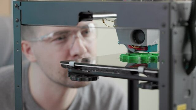engineer man wearing glasses watching the process of printing the gear detail from green plastic in a 3d printer.