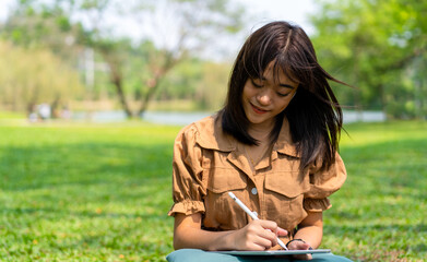 happy young Asian female student spends holiday time writing journal, planning future things and studying for school classes, sitting on grass at public park in relaxed greenery environment