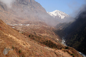 Valle de Langtang, con un estrecho río entre montañas, un poblado y un pico nevado al fondo, con vegetación seca y nubes bajas