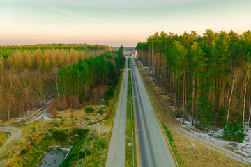 Asfaltowa droga przez sosnowy, wysoki las. Z lewej strony widać latarnie uliczne i chodnik ze ścieżką rowerową. Widok z wysokości, zdjęcie zrobione z użyciem drona.