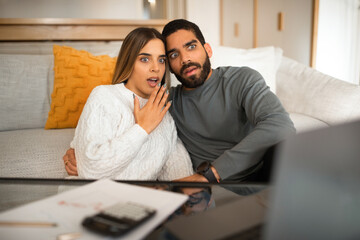 Scared shocked international millennial couple looking at laptop, have video call, pay taxes in...