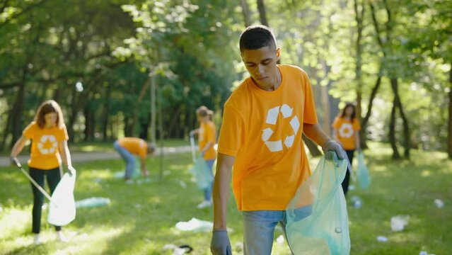 Multiethnic Young Man Using Trash Picker While Collecting Garbage in Bin Bag. Group of Diverse Volunteers in T-Shirts with Recycling Symbol Cleaning Public Park. Safe Ecology Concept