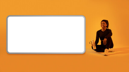 Workout app. Black lady sitting near huge cellphone screen, resting after training on orange neon background, mockup