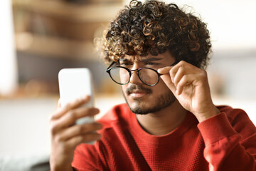 Eyesight Problems Concept. Young Indian Guy Wearing Eyeglasses Reading Message On Smartphone