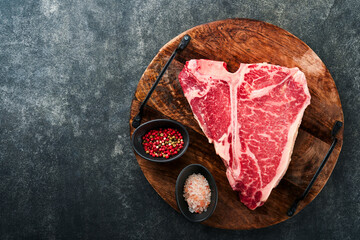 Thick Raw T-Bone Steak. Dry-aged Raw T-bone or porterhouse beef meat Steak on cutting boar with herbs and salt on dark background. Top view and copy space.