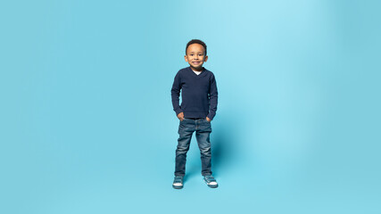 Naklejka premium Happy childhood. Full length shot of black little boy holding hands in pockets, blue background, panorama, free space