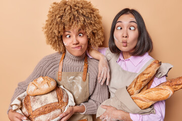 Two housewifes bake bread at home make funny grimaces have face dirty with flour hold round freshly baked bread and baguettes wear aprons involved in domesticc production isolated on brown background - obrazy, fototapety, plakaty