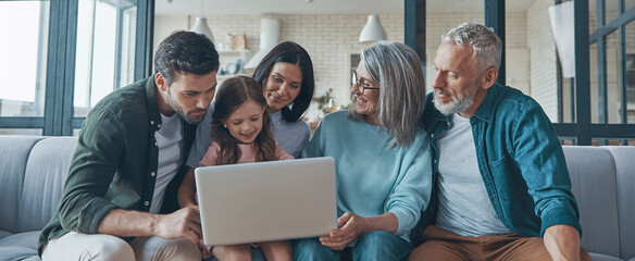 Happy multi-generation family using laptop and smiling while spending time home together - Powered by Adobe