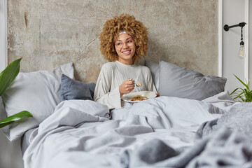 Pleased beautiful woman enjoys breakfast in bed during morning looks happily aside eats muesli...