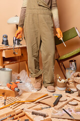 Faceless female carpenter dressed in brown overalls boots and protective gloves holds instruments for woodworking poses in workshop assembles wooden furniture uses carpentry tools. Cropped shot