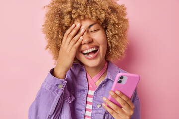 Cheerful curly haired woman makes face palm smiles broadly shows white teeth holdds smartphone reads funny content dressed in fashionable purple jacket isolated over pink background. Positive emotions