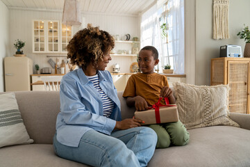 African American child son presenting surprised happy mom with gift while celebrating Mothers Day together at home. Smiling excited mother receiving birthday present from kid