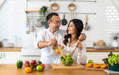 Young asian family couple having fun cooking and preparing cook vegan food healthy eat with fresh vegetable salad on counter in kitchen at home.Happy couple looking to preparing food